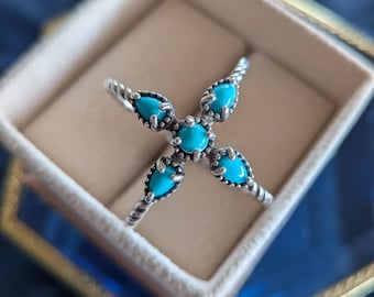 Könnte beinhalten: Ein silberner Ring mit Kreuzdesign, besetzt mit fünf leuchtend türkisfarbenen, tropfenförmigen Steinen. Der Ring wird in einer beigen Schmuckschatulle mit goldverziertem Glasdeckel vor dunklem blauem Hintergrund präsentiert.