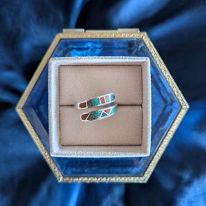 May include: A silver ring with a wrap-around design, featuring inlaid sections of red, green, and blue mosaic-style stones. The ring is displayed in a hexagonal glass jewelry box with a gold-coloured frame, set against a dark blue background.