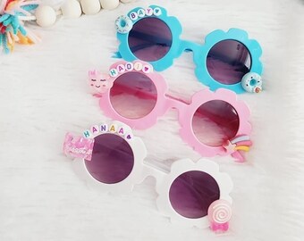 May include: Three pairs of colorful flower-shaped sunglasses for kids. The sunglasses have different colored frames and lenses, and each pair has a different cute decoration on the side. The decorations include a donut, a unicorn, and a rainbow.