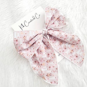 May include: A large, light pink hair bow with a white stitched edge. The bow features a repeating pattern of ballerinas, trees, and stars. A white card with the text "Kb Created Co." is tucked under the bow.