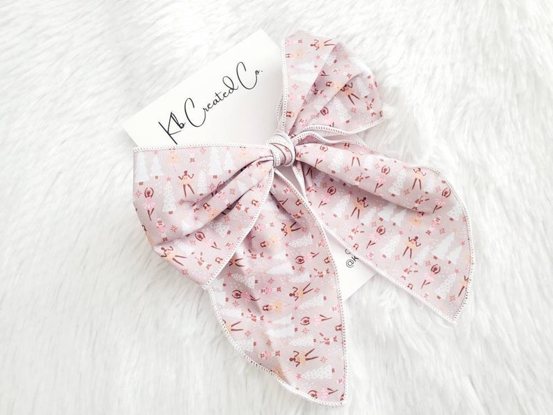 May include: A large, light pink hair bow with a white stitched edge. The bow features a repeating pattern of ballerinas, trees, and stars. A white card with the text "Kb Created Co." is tucked under the bow.