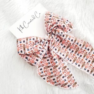May include: A large, decorative hair bow featuring a repeating pattern of toy soldiers in shades of pink, brown, and black on a white background. The bow is tied in a large knot with long, flowing tails. The text "Kb Created Co." is visible on a white card.