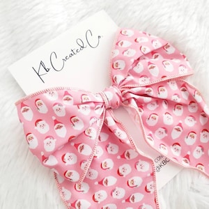 May include: A large pink bow with a repeating pattern of Santa Claus faces. The bow is tied in the center and has long, flowing tails. A white card with the text "Kb Created Co." is in the background.