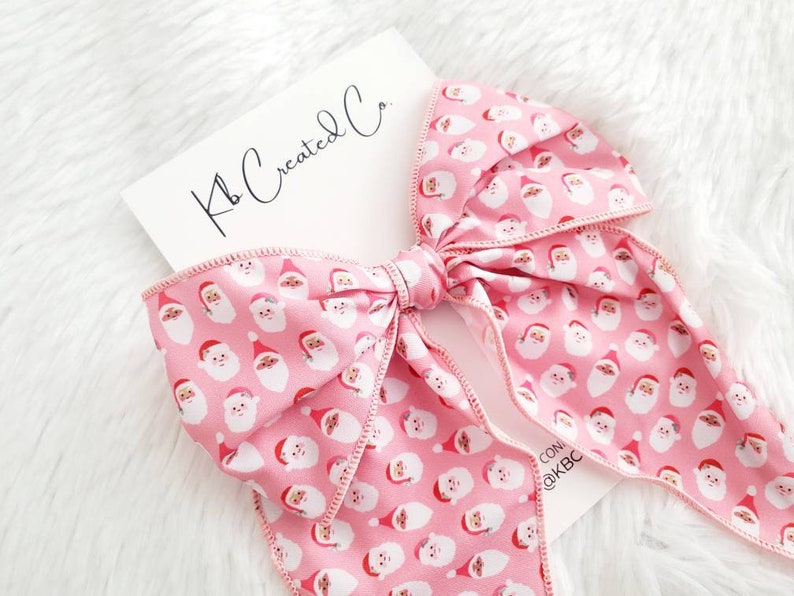 May include: A large pink bow with a repeating pattern of Santa Claus faces. The bow is tied in the center and has long, flowing tails. A white card with the text "Kb Created Co." is in the background.