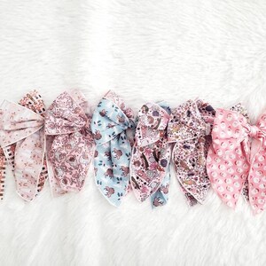 May include: A collection of seven fabric hair bows in various patterns and colors. The bows feature floral, geometric, and whimsical designs in shades of pink, blue, and brown. The bows are arranged on a white, fluffy surface.