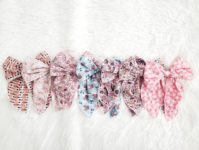 May include: A collection of seven fabric hair bows in various patterns and colors. The bows feature floral, geometric, and whimsical designs in shades of pink, blue, and brown. The bows are arranged on a white, fluffy surface.