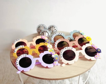 May include: A set of six colorful flower-shaped sunglasses with personalized names, including "Kleo Minnie", "Brooklyn", "Celia", "Anastasia", and two others. The sunglasses are on a wooden table with a white background.