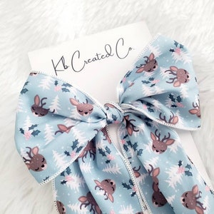 May include: A light blue hair bow with a repeating pattern of deer, holly, and Christmas trees. The bow has a white stitched edge and is attached to a white card with the text "Kb Created Co."