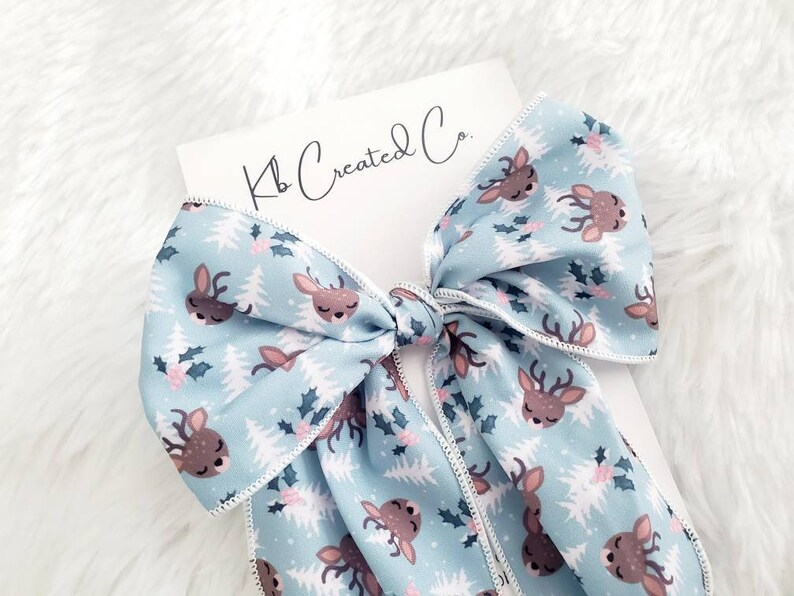 May include: A light blue hair bow with a repeating pattern of deer, holly, and Christmas trees. The bow has a white stitched edge and is attached to a white card with the text "Kb Created Co."