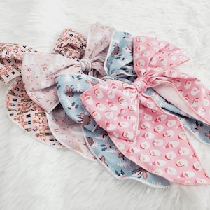 May include: A collection of Christmas-themed hair bows. The bows feature various patterns, including Santa Claus faces, reindeer, and festive geometric designs. The color palette includes pink, blue, and neutral tones, creating a cheerful holiday aesthetic.