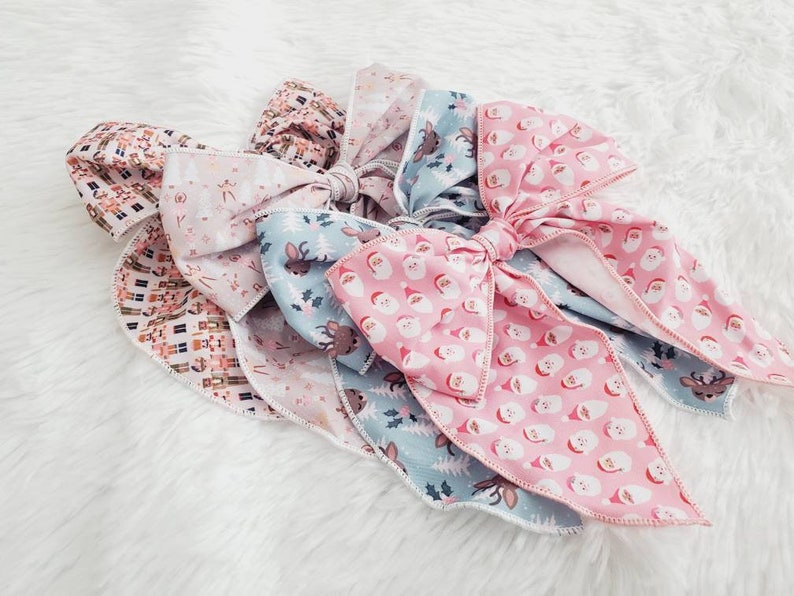 May include: A collection of Christmas-themed hair bows. The bows feature various patterns, including Santa Claus faces, reindeer, and festive geometric designs. The color palette includes pink, blue, and neutral tones, creating a cheerful holiday aesthetic.