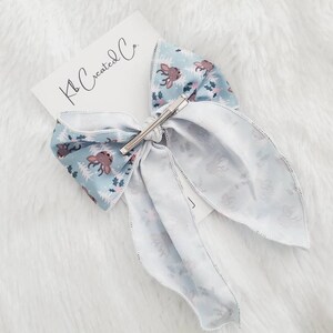May include: A light blue hair bow with a reindeer and tree pattern, secured with a silver barrette. The bow is attached to a white card with the text "Kb Created Co."