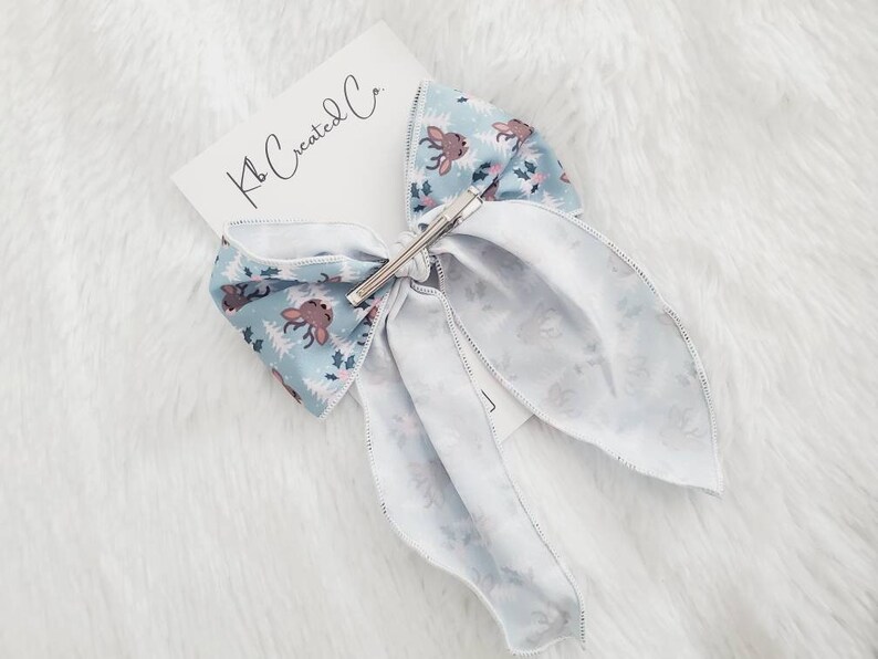 May include: A light blue hair bow with a reindeer and tree pattern, secured with a silver barrette. The bow is attached to a white card with the text "Kb Created Co."