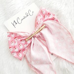 May include: A pink hair bow with a gold barrette clip. The bow features a pattern of Santa Claus faces on a light pink background. The text "Kb Created Co." is on a white card.