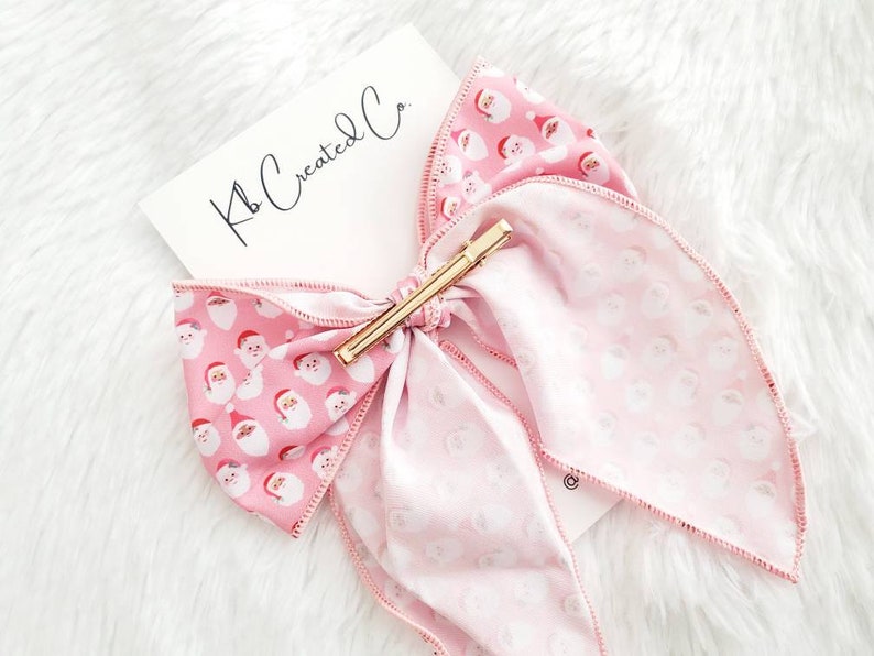 May include: A pink hair bow with a gold barrette clip. The bow features a pattern of Santa Claus faces on a light pink background. The text "Kb Created Co." is on a white card.