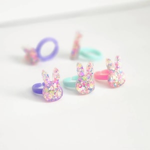 Puede incluir: Cinco anillos de conejito de purpurina de colores. Los anillos son de plástico y tienen un acabado brillante. Los conejitos son en tonos de rosa, morado y azul.