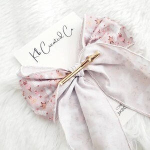 May include: A light purple hair bow with a gold clip. The bow features a floral pattern with pink and white accents. The tag reads "Kb Created Co."