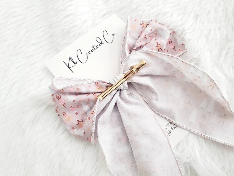 May include: A light purple hair bow with a gold clip. The bow features a floral pattern with pink and white accents. The tag reads "Kb Created Co."
