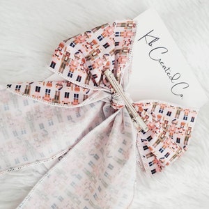 May include: A large, decorative hair bow with a repeating pattern of toy soldiers in shades of brown, pink, and blue on a white background. The bow is secured with a silver metal clip. A white card with the text "Kb Created Co." is visible.
