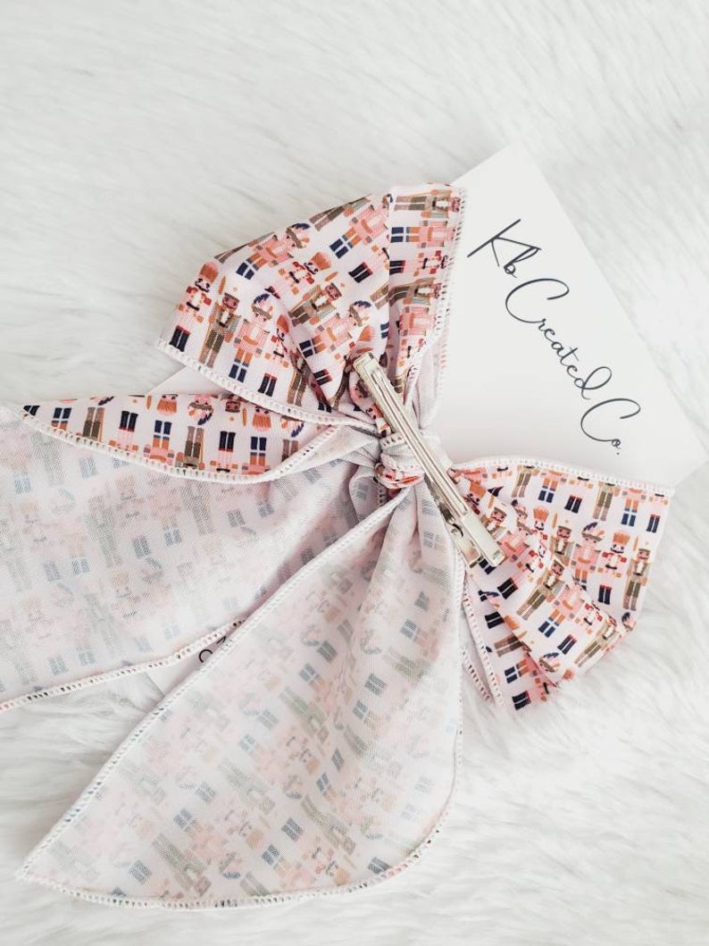 May include: A large, decorative hair bow with a repeating pattern of toy soldiers in shades of brown, pink, and blue on a white background. The bow is secured with a silver metal clip. A white card with the text "Kb Created Co." is visible.