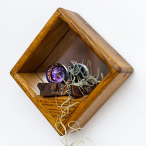Può includere: Una mensola da parete in legno a forma di rombo con specchio sul retro, contenente una piccola sfera di vetro iridescente con macchie viola e dorate, poggiata su un pezzo di legno scuro e decorata con una pianta aerea ricadente.