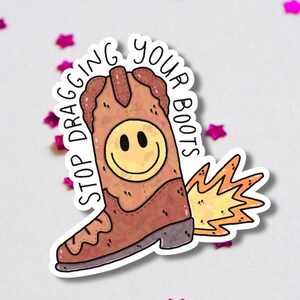 Pode incluir: Um autocolante com uma bota de cowboy castanha com um smiley amarelo e o texto "STOP DRAGGING YOUR BOOTS". A bota tem um desenho de chama e é rodeada por confetes de estrelas cor-de-rosa.