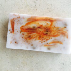 May include: A rectangular bar of handmade soap with a white base and orange and brown swirls. The soap has a speckled appearance with small brown dots throughout. The design resembles a stylized flower or abstract pattern.