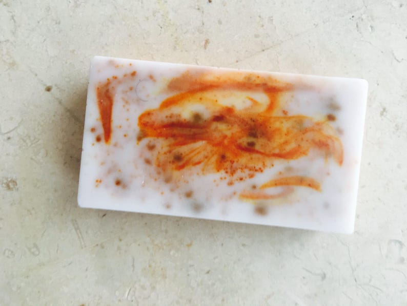 May include: A rectangular bar of handmade soap with a white base and orange and brown swirls. The soap has a speckled appearance with small brown dots throughout. The design resembles a stylized flower or abstract pattern.