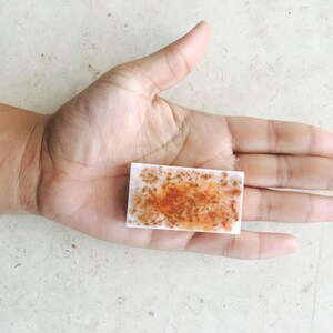 May include: A rectangular, white soap bar with a mottled orange and brown pattern. The soap is held in an open hand, showcasing its size and design. The soap appears to be handmade.