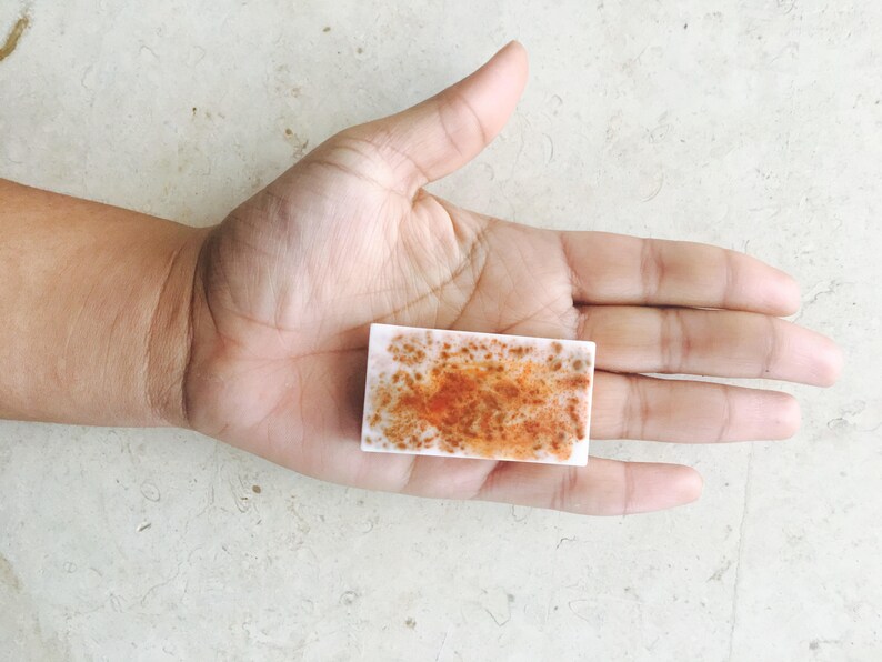 May include: A rectangular, white soap bar with a mottled orange and brown pattern. The soap is held in an open hand, showcasing its size and design. The soap appears to be handmade.