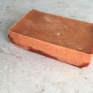May include: A rectangular bar of handmade soap. The soap has a warm, orange-brown hue with subtle speckles and a marbled appearance. The edges show a darker, reddish-brown colour. The soap is likely scented and suitable for personal care.