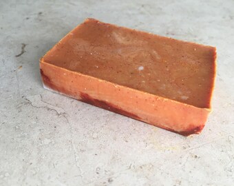 May include: A rectangular bar of handmade soap. The soap has a warm, orange-brown hue with subtle speckles and a marbled appearance. The edges show a darker, reddish-brown colour. The soap is likely scented and suitable for personal care.