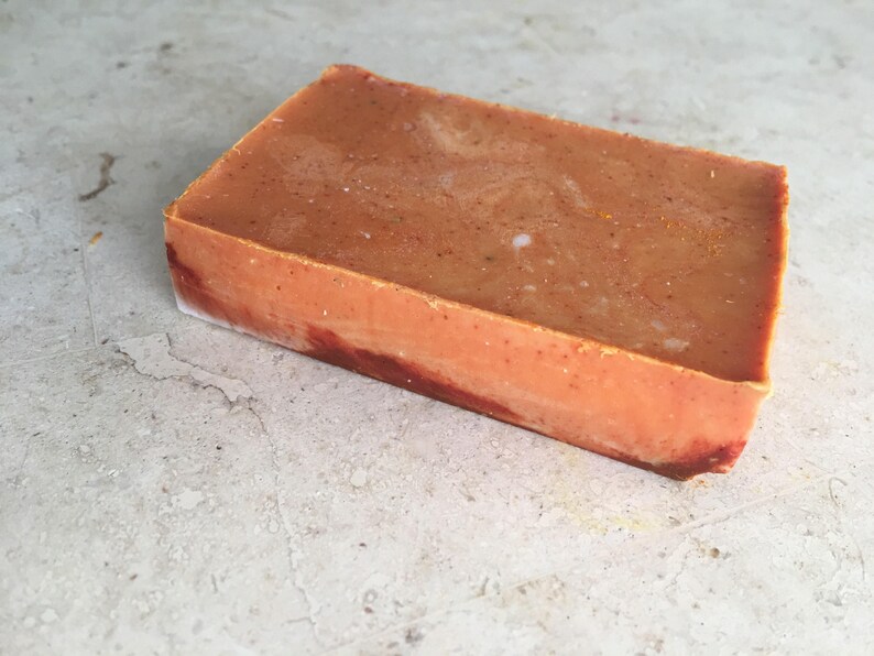 May include: A rectangular bar of handmade soap. The soap has a warm, orange-brown hue with subtle speckles and a marbled appearance. The edges show a darker, reddish-brown colour. The soap is likely scented and suitable for personal care.