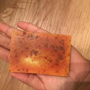 May include: A rectangular bar of soap with a mottled orange and brown surface. The soap is held in a hand, and the background is a wooden surface. The soap has a light-colored center.
