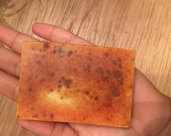 May include: A rectangular bar of soap with a mottled orange and brown surface. The soap is held in a hand, and the background is a wooden surface. The soap has a light-coloured centre.