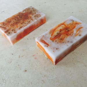 May include: Two rectangular bars of handmade soap. The soap has a white top layer with orange and brown accents. The sides are orange. One bar has a speckled design, and the other has an orange floral design.