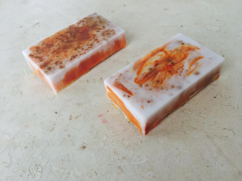 May include: Two rectangular bars of handmade soap. The soap has a white top layer with orange and brown accents. The sides are orange. One bar has a speckled design, and the other has an orange floral design.