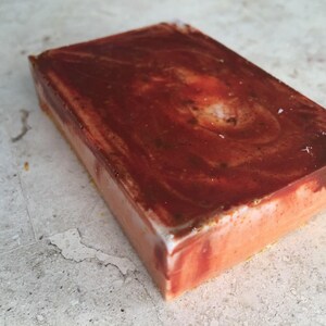 May include: A rectangular bar of handmade soap with a marbled design. The top layer is a deep reddish-brown, swirling into lighter shades of red and orange. The sides are a pale orange with a white edge.