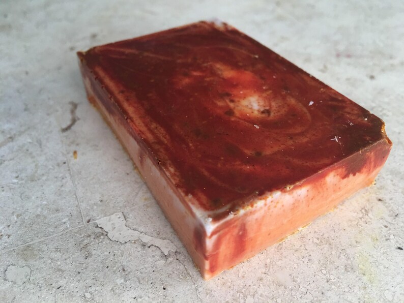 May include: A rectangular bar of handmade soap with a marbled design. The top layer is a deep reddish-brown, swirling into lighter shades of red and orange. The sides are a pale orange with a white edge.