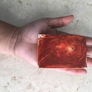 May include: A rectangular bar of soap with a reddish-brown, swirled pattern. The soap is held in a hand, demonstrating its size and texture. The soap appears handmade, with a natural aesthetic.
