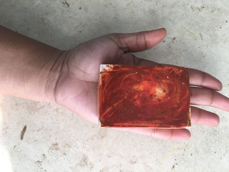 May include: A rectangular bar of soap with a reddish-brown, swirled pattern. The soap is held in a hand, demonstrating its size and texture. The soap appears handmade, with a natural aesthetic.