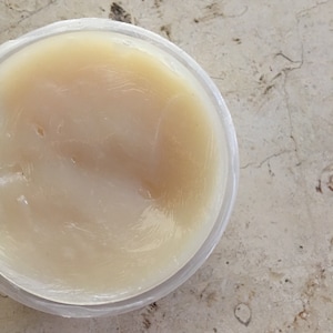 May include: A clear, round plastic container filled with a light yellow, semi-solid substance. The substance has a smooth, glossy appearance. The container sits on a light-colored, speckled surface.