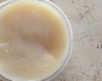May include: A clear, round plastic container filled with a pale yellow, semi-solid substance. The substance has a smooth, glossy appearance. The container sits on a light-coloured, speckled surface.