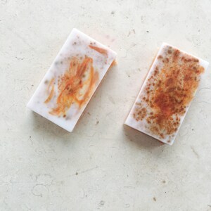 May include: Two rectangular bars of handmade soap. The soap has a white base with orange and brown marbling. The bars are displayed on a light-colored, textured surface.