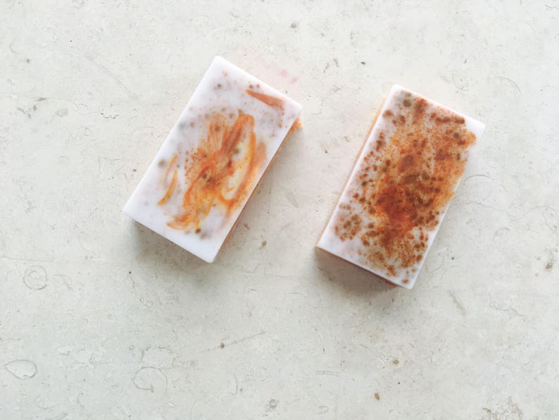 May include: Two rectangular bars of handmade soap. The soap has a white base with orange and brown marbling. The bars are displayed on a light-colored, textured surface.