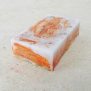 May include: A rectangular bar of handmade soap with a white top and orange swirls. The soap has a floral design in the center and is set against a neutral background. The soap appears to be scented.