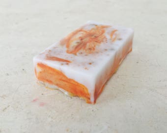 May include: A rectangular bar of handmade soap with a white top and orange swirls. The soap has a floral design in the center and is set against a neutral background. The soap appears to be scented.