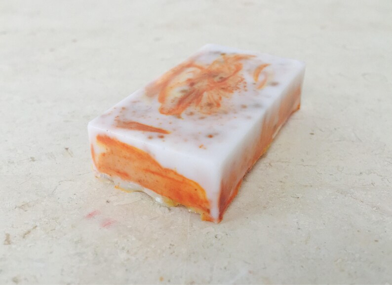 May include: A rectangular bar of handmade soap with a white top and orange swirls. The soap has a floral design in the center and is set against a neutral background. The soap appears to be scented.