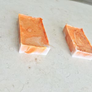 May include: Two rectangular bars of handmade soap. The soap has a marbled orange and white design. The bars are angled, with the orange color on top and white on the bottom. The soap is on a light-colored surface.