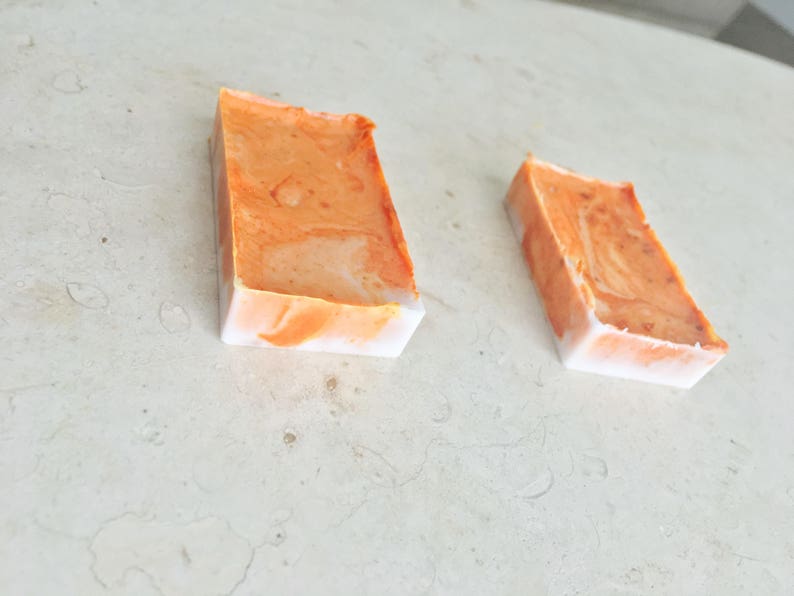 May include: Two rectangular bars of handmade soap. The soap has a marbled orange and white design. The bars are angled, with the orange color on top and white on the bottom. The soap is on a light-colored surface.